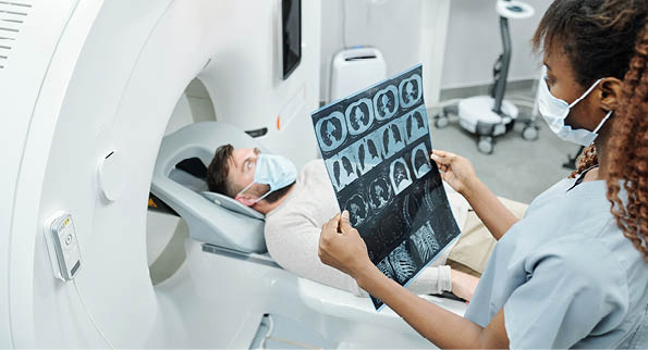 Young African radiologist in uniform and protective mask looking at x-ray image of patient lying on long couchette of medical equipment